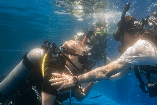 Three scuba divers in a swimming pool, with one PADI Instructor Course Trainee appearing to instruct the others. They wear wetsuits, masks, and air tanks, surrounded by clear water with light reflections.