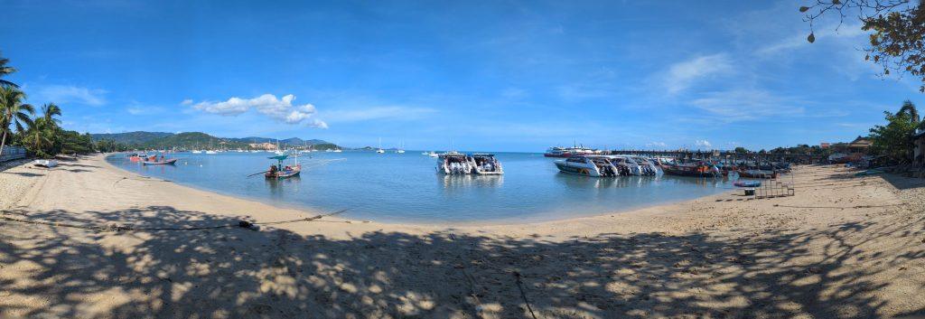 A serene beach with golden sand and calm waters under a clear blue sky. Several boats are anchored near the shore. Hills and lush greenery are visible in the distance, creating a picturesque backdrop.