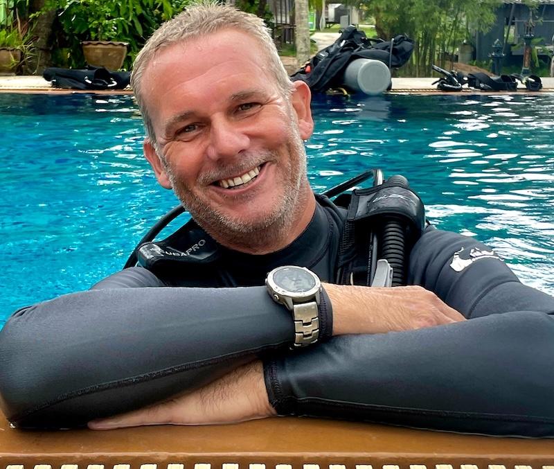 A man in a wetsuit smiles while leaning on the edge of a pool. He is wearing a dive watch, and scuba gear is visible behind him. The water is a bright blue, and lush greenery surrounds the pool area.