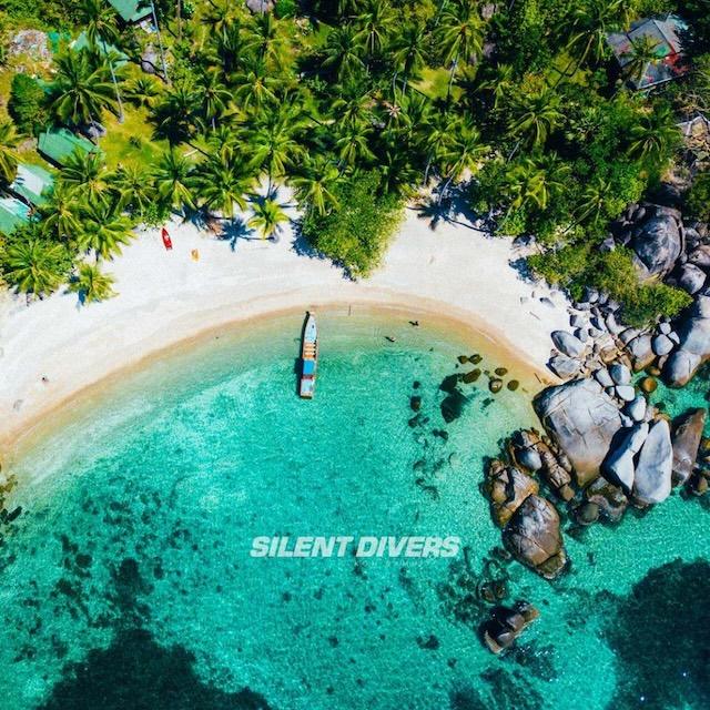 Aerial view of a tropical beach with clear turquoise water, a small boat near the shore, lush green palm trees, and large rocks scattered along the coastline. The words "Silent Divers" are visible on the image.